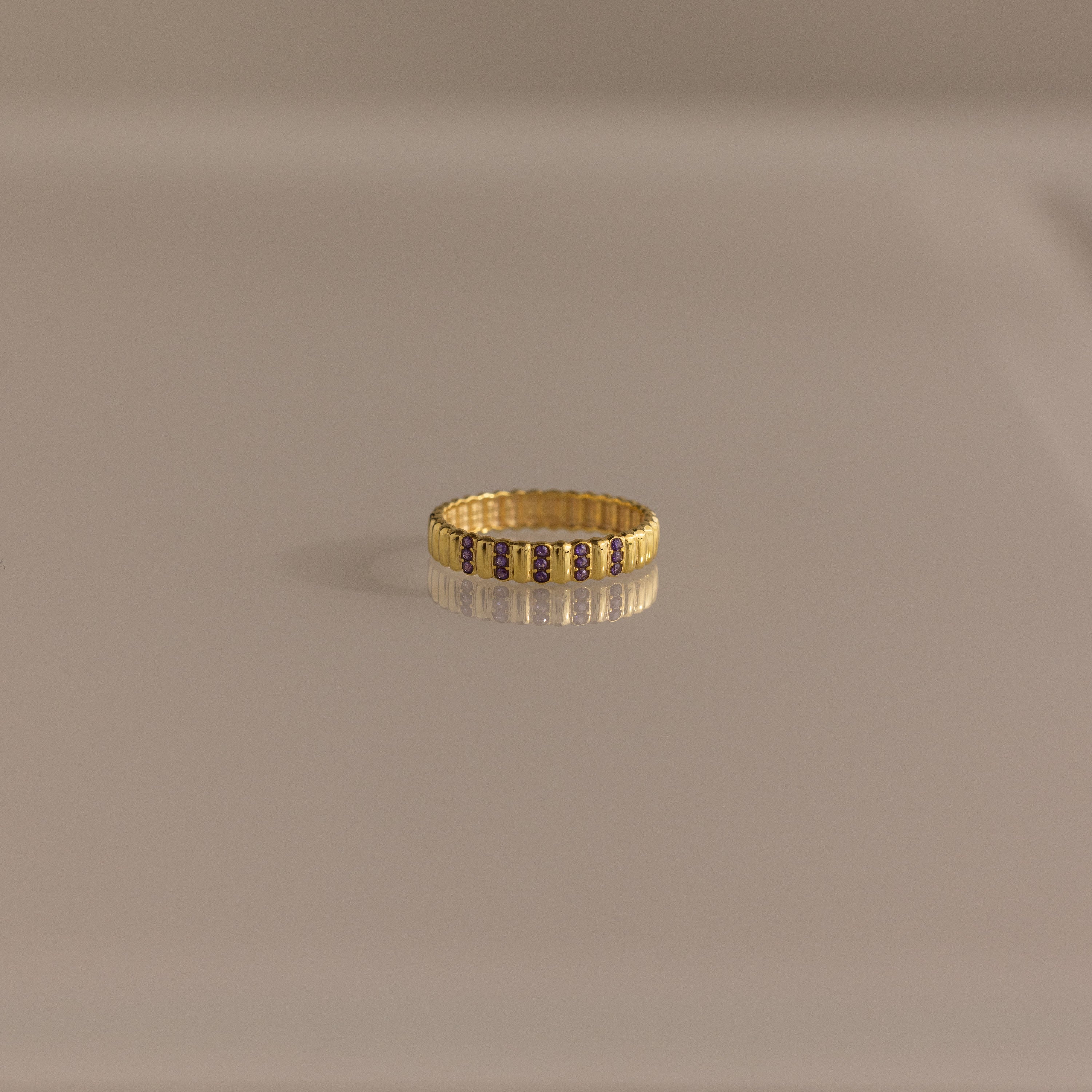 The Ribbed Triple Alexandrite Ring, featuring small, evenly spaced alexandrite stones, is displayed on a reflective beige surface.