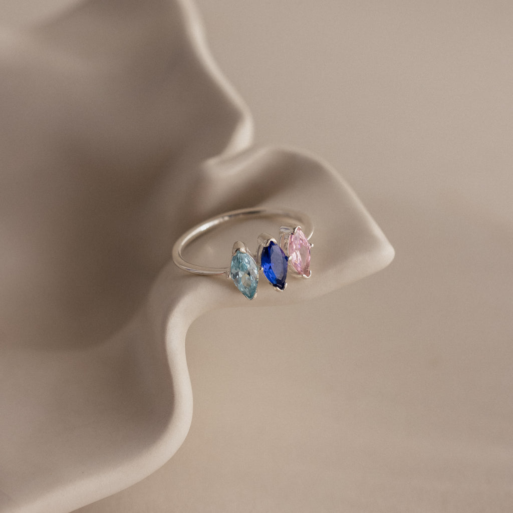 The Multiple Marquise Birthstone Ring showcases three marquise-cut stones in blue, navy, and pink, elegantly displayed on a wavy beige surface.