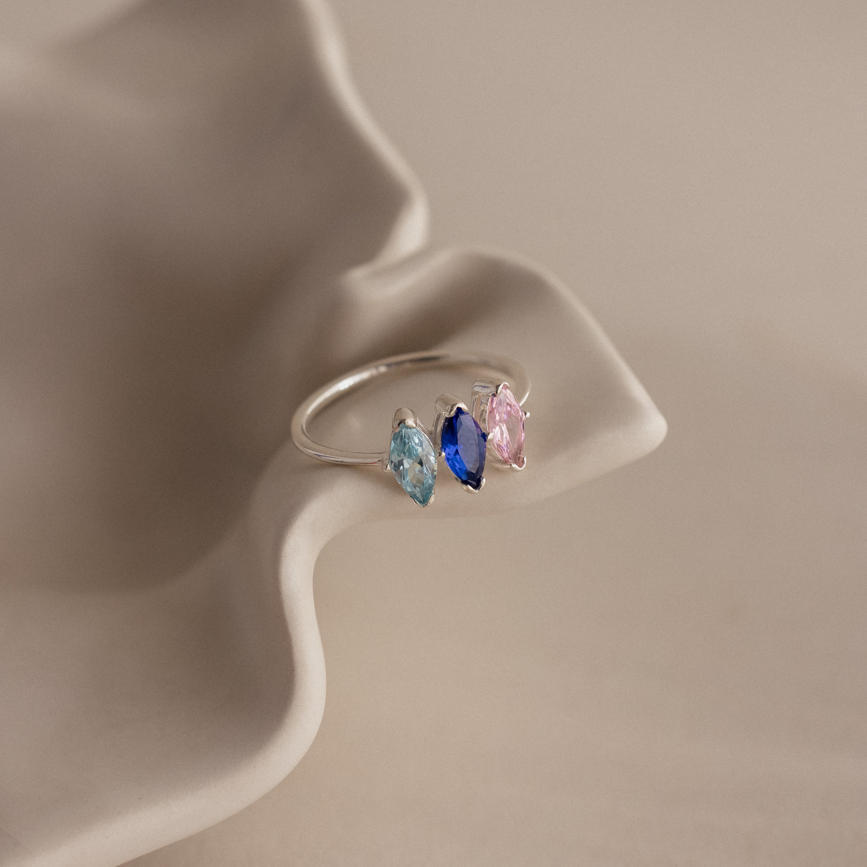 The Multiple Marquise Birthstone Ring showcases three marquise-cut stones in blue, navy, and pink, elegantly displayed on a wavy beige surface.