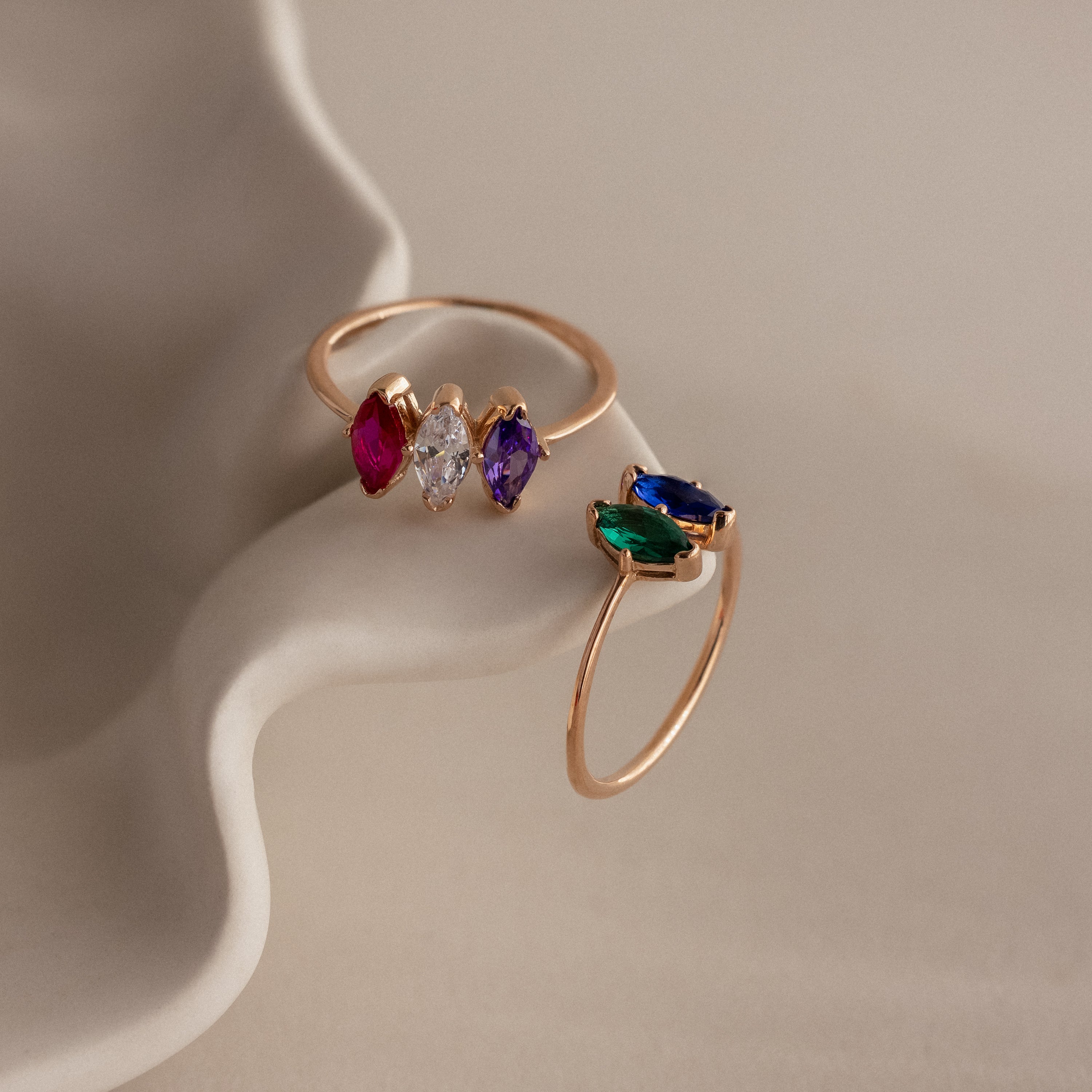Two Multiple Marquise Birthstone Rings in Rose Gold, adorned with vibrant gemstones, rest on a wavy cream surface—ideal as personalized jewelry or a meaningful milestone gift.