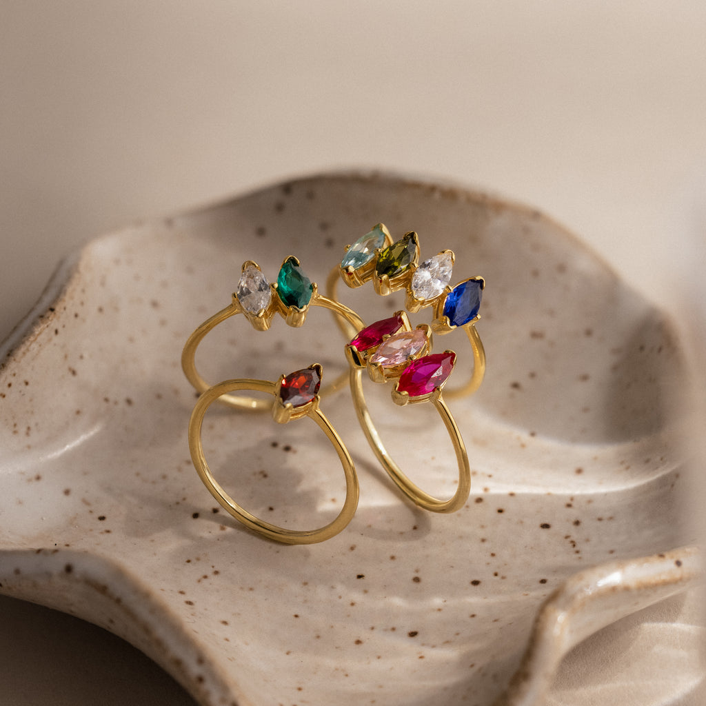 The Multiple Marquise Birthstone Rings in 18K Gold, including a personalized birthstone option, are displayed on a speckled ceramic dish.