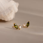 Marquise Peridot & Pearl Studs, featuring green marquise stones and white pearls in a toi et moi design, are showcased on a beige surface beside a seashell.