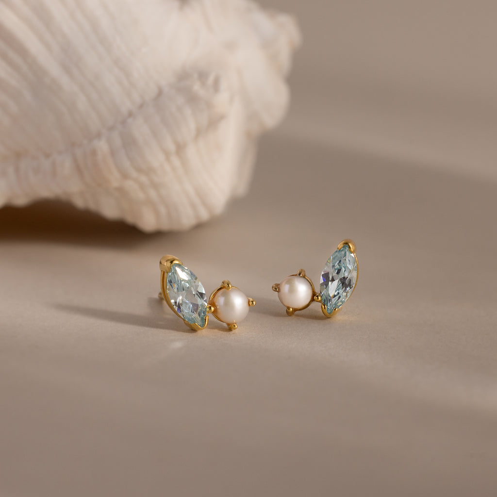 Marquise Aquamarine & Pearl Studs are elegantly displayed on a beige surface beside a white seashell.