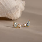 Marquise Aquamarine & Pearl Studs are elegantly displayed on a beige surface beside a white seashell.