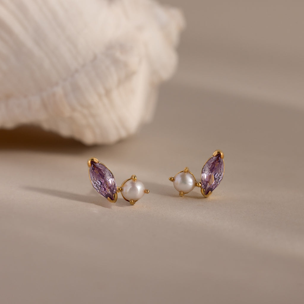 Marquise Alexandrite & Pearl Studs, featuring gold toi et moi design with purple gemstones and pearls, displayed on a neutral surface with a shell in the background.