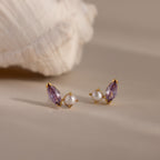 Marquise Alexandrite & Pearl Studs, featuring gold toi et moi design with purple gemstones and pearls, displayed on a neutral surface with a shell in the background.