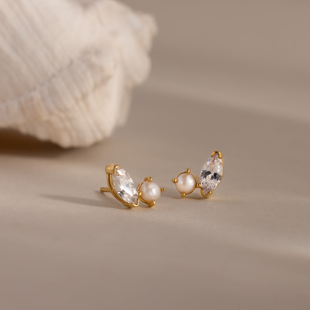 Marquise Opal & Pearl Studs—gold earrings featuring pearl and marquise opal details in a toi et moi design—are displayed on a neutral surface next to a seashell.