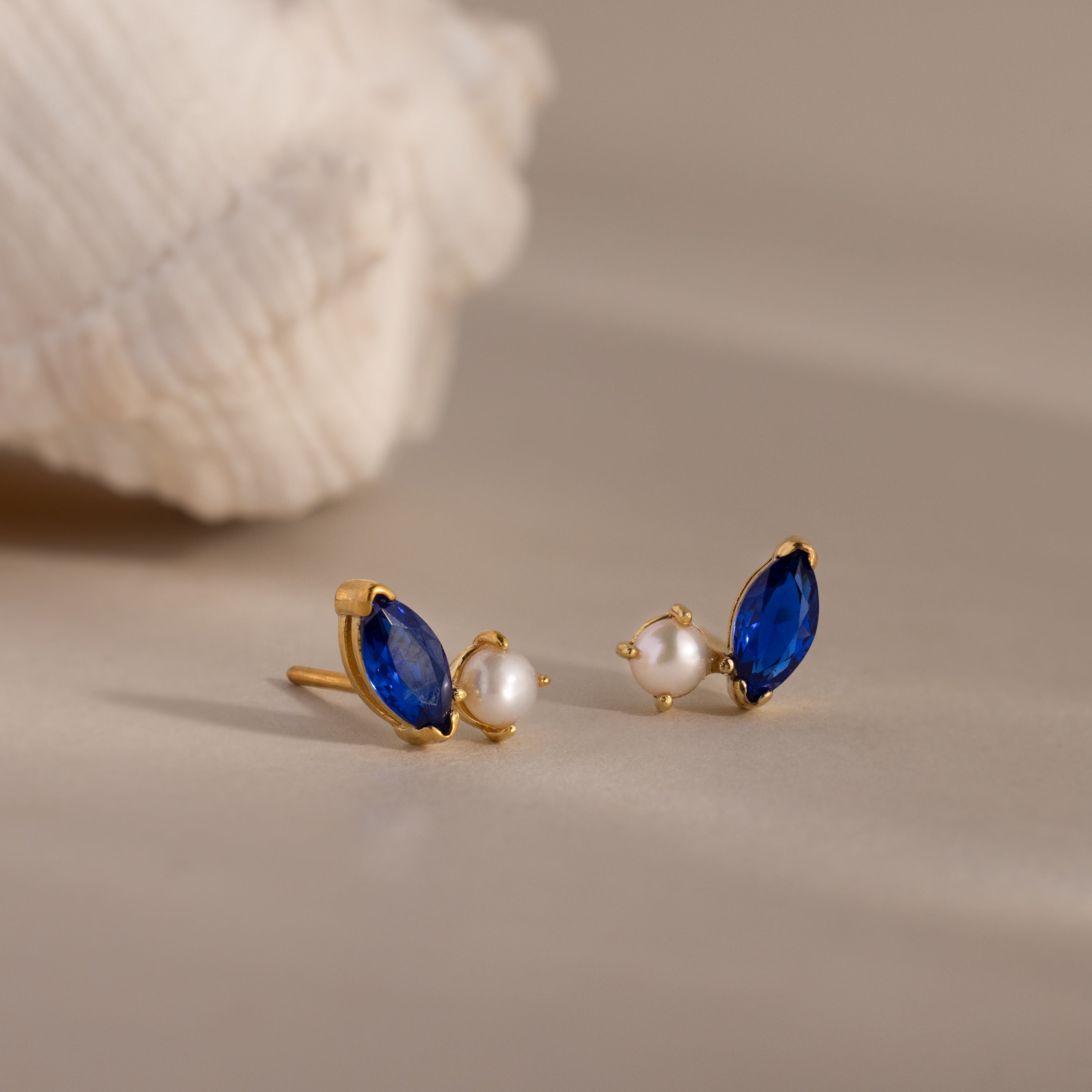 Marquise Sapphire & Pearl Studs—gold earrings with blue marquise stones and small pearls—are displayed on a beige surface beside a seashell.