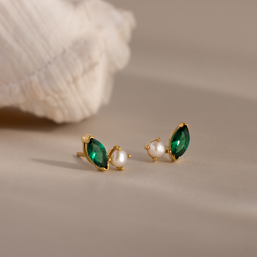 Gold earrings with green marquise stones and pearls rest on a beige surface, accented by a shell—elegant Marquise Emerald & Pearl Studs for a timeless look.