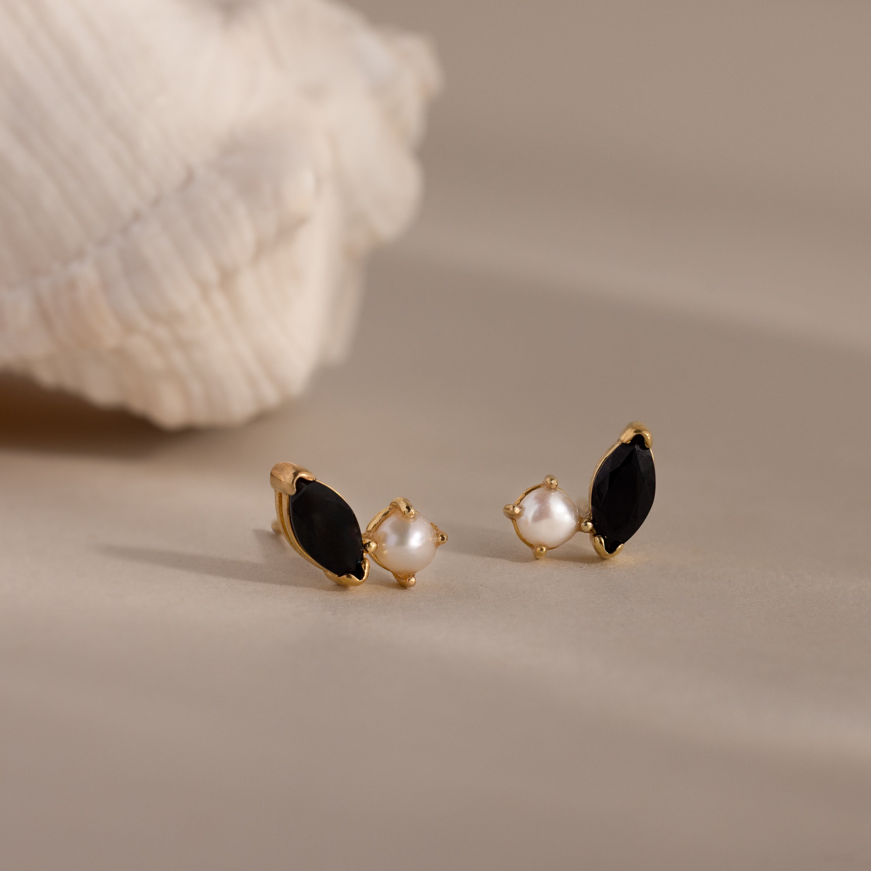 Marquise Onyx & Pearl Studs—gold earrings featuring black stones and pearls in an elegant toi et moi design—displayed on a neutral surface beside a white textured shell.