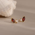 Marquise Garnet & Pearl Studs, featuring gold earrings with red garnet gemstones and pearls inspired by marquise birthstone designs, rest on a beige surface with a shell in the background.