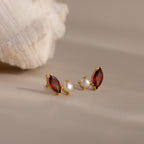 Marquise Garnet & Pearl Studs, featuring gold earrings with red garnet gemstones and pearls inspired by marquise birthstone designs, rest on a beige surface with a shell in the background.