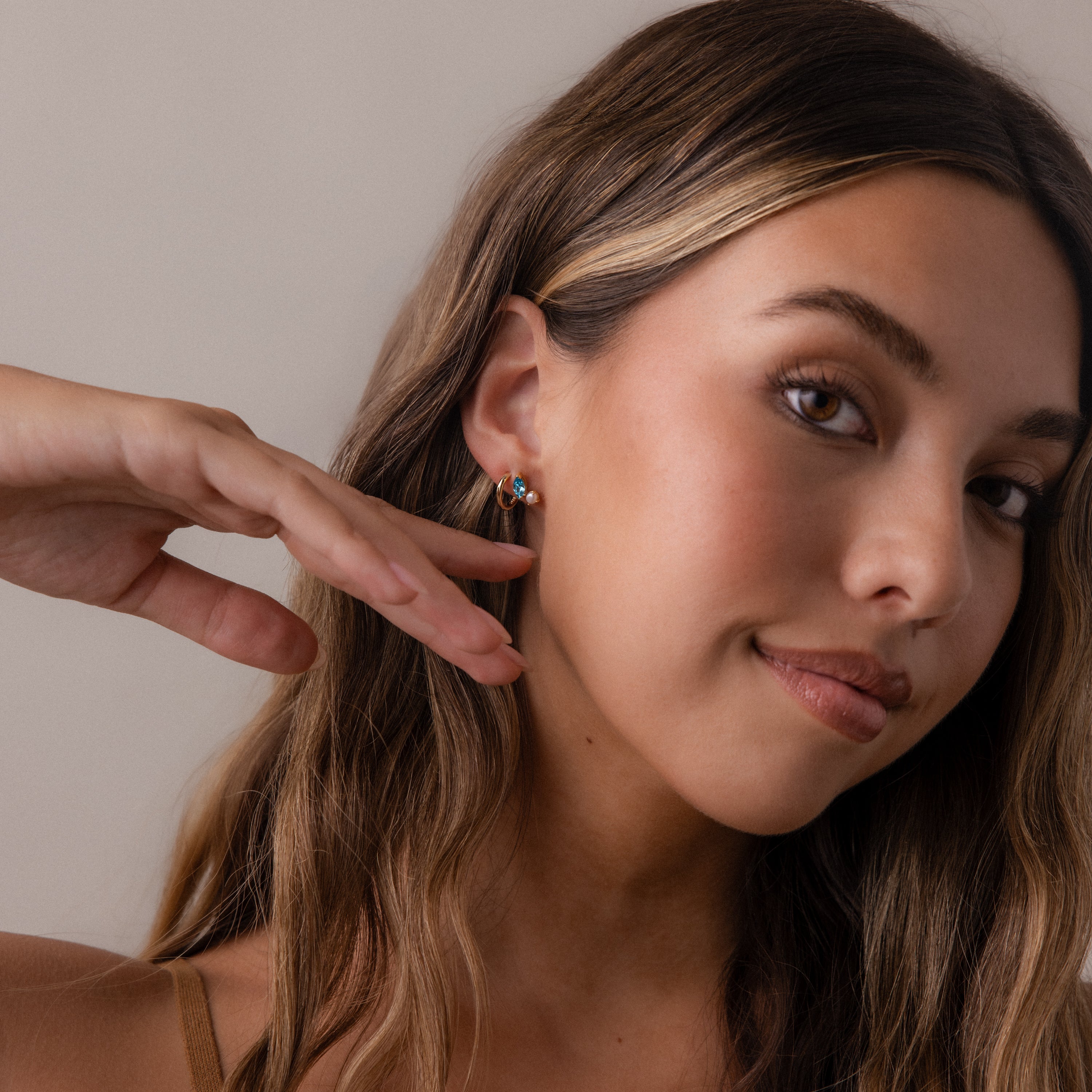 A woman with long brown hair and subtle makeup touches her Marquise Birthstone & Pearl Studs while posing against a neutral background.
