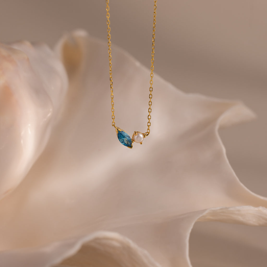 The Marquise Zircon & Pearl Necklace, featuring a gold chain and blue marquise pendant, is elegantly displayed before a large white seashell.