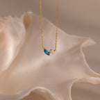 The Marquise Zircon & Pearl Necklace, featuring a gold chain and blue marquise pendant, is elegantly displayed before a large white seashell.