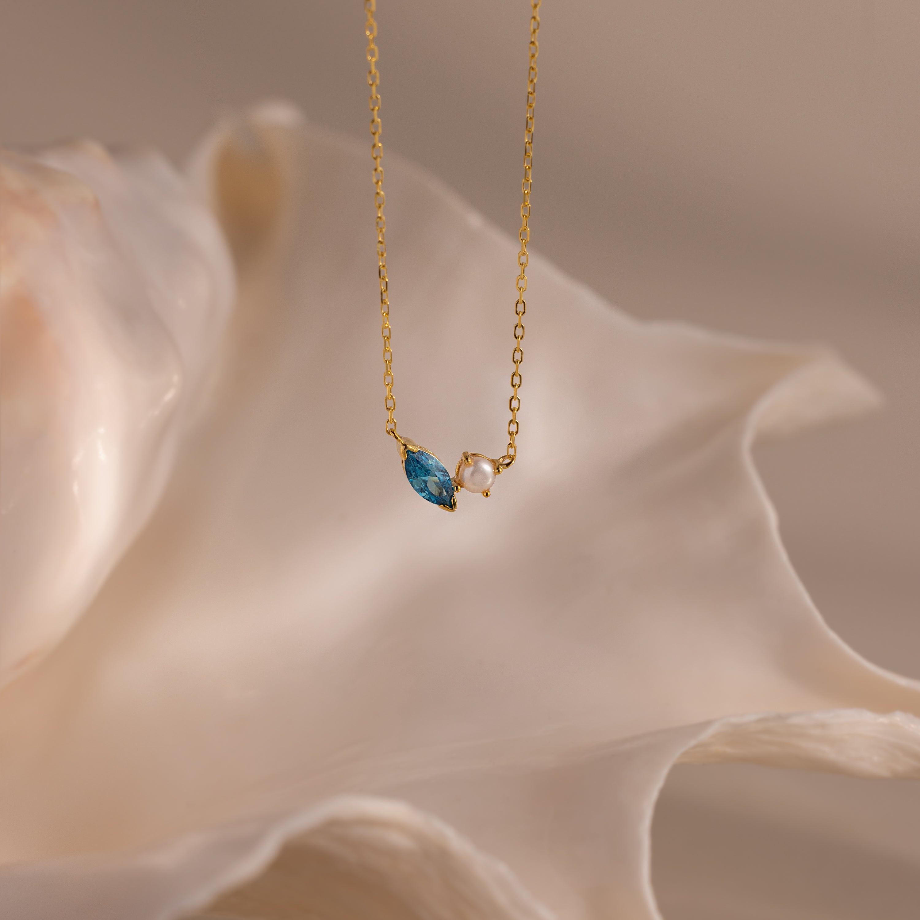 The Marquise Zircon & Pearl Necklace, featuring a gold chain and blue marquise pendant, is elegantly displayed before a large white seashell.