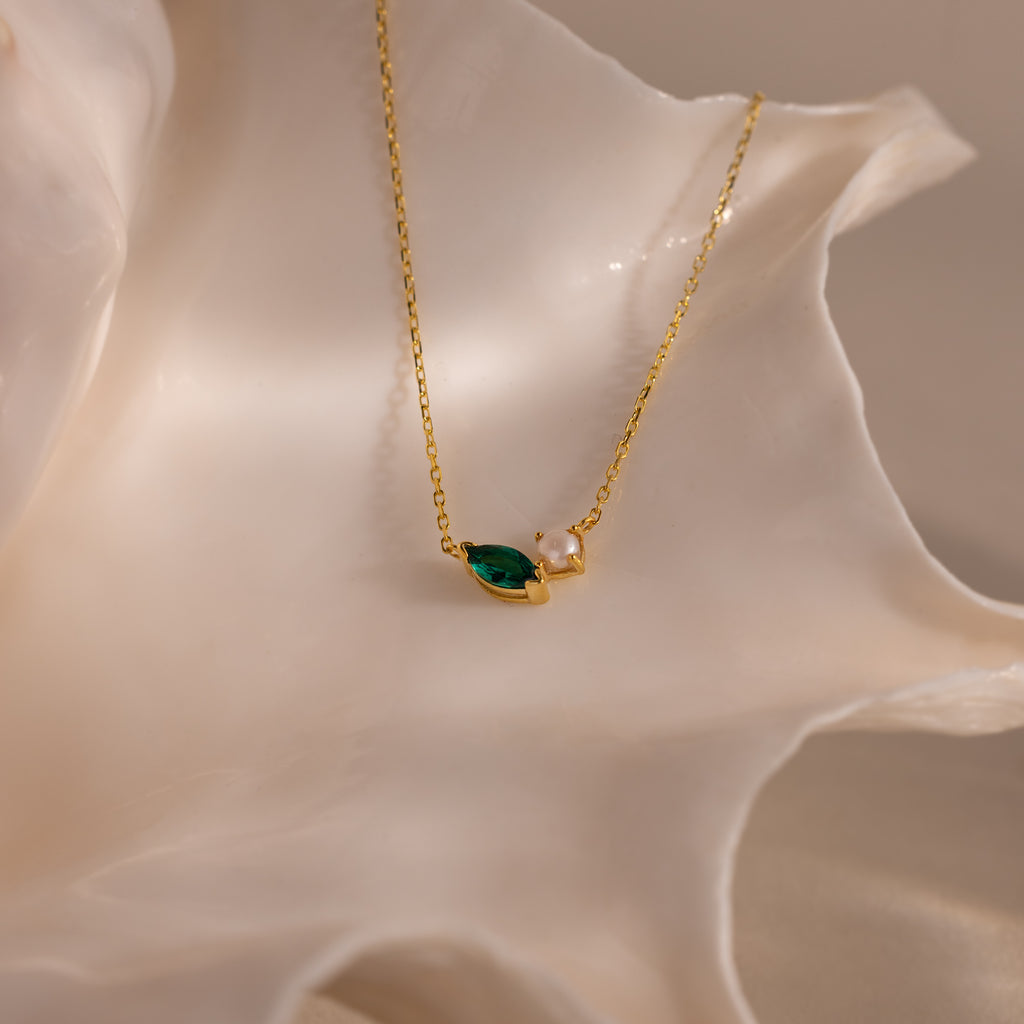 The Marquise Emerald & Pearl Necklace, featuring a green marquise birthstone and a small pearl, is displayed on a white seashell backdrop.