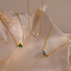 The Marquise Birthstone & Pearl Necklace, along with two other gold jeweled pendant necklaces, are elegantly displayed on a large, glossy seashell.