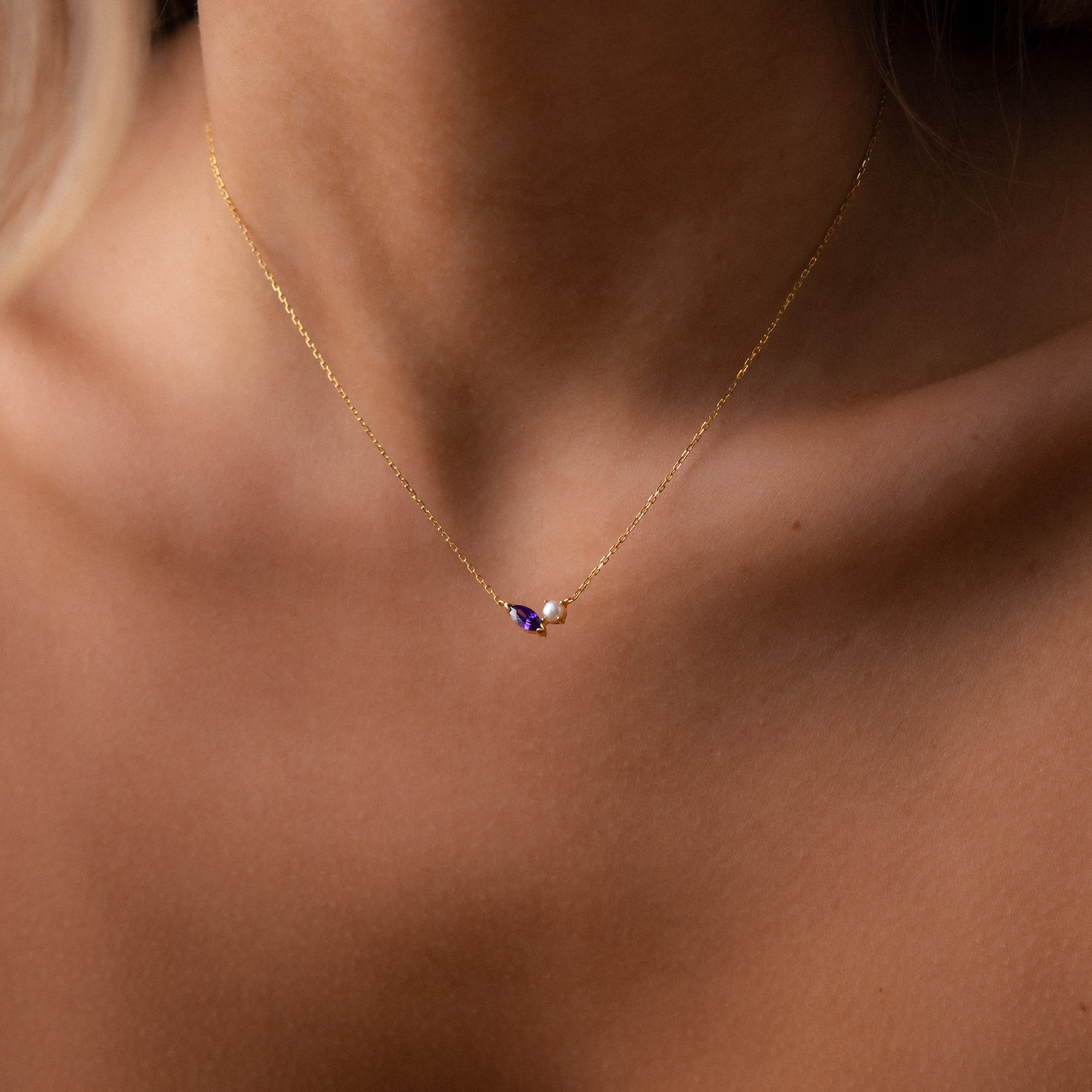The Marquise Zircon & Pearl Necklace features a gold chain with a purple marquise zircon birthstone and pearl pendant, elegantly draped on a woman's neck.