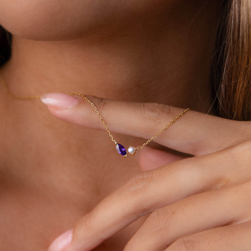 Close-up of a hand holding the Marquise Birthstone & Pearl Necklace, a gold customizable piece featuring a purple gem and a small pearl, shown against skin.