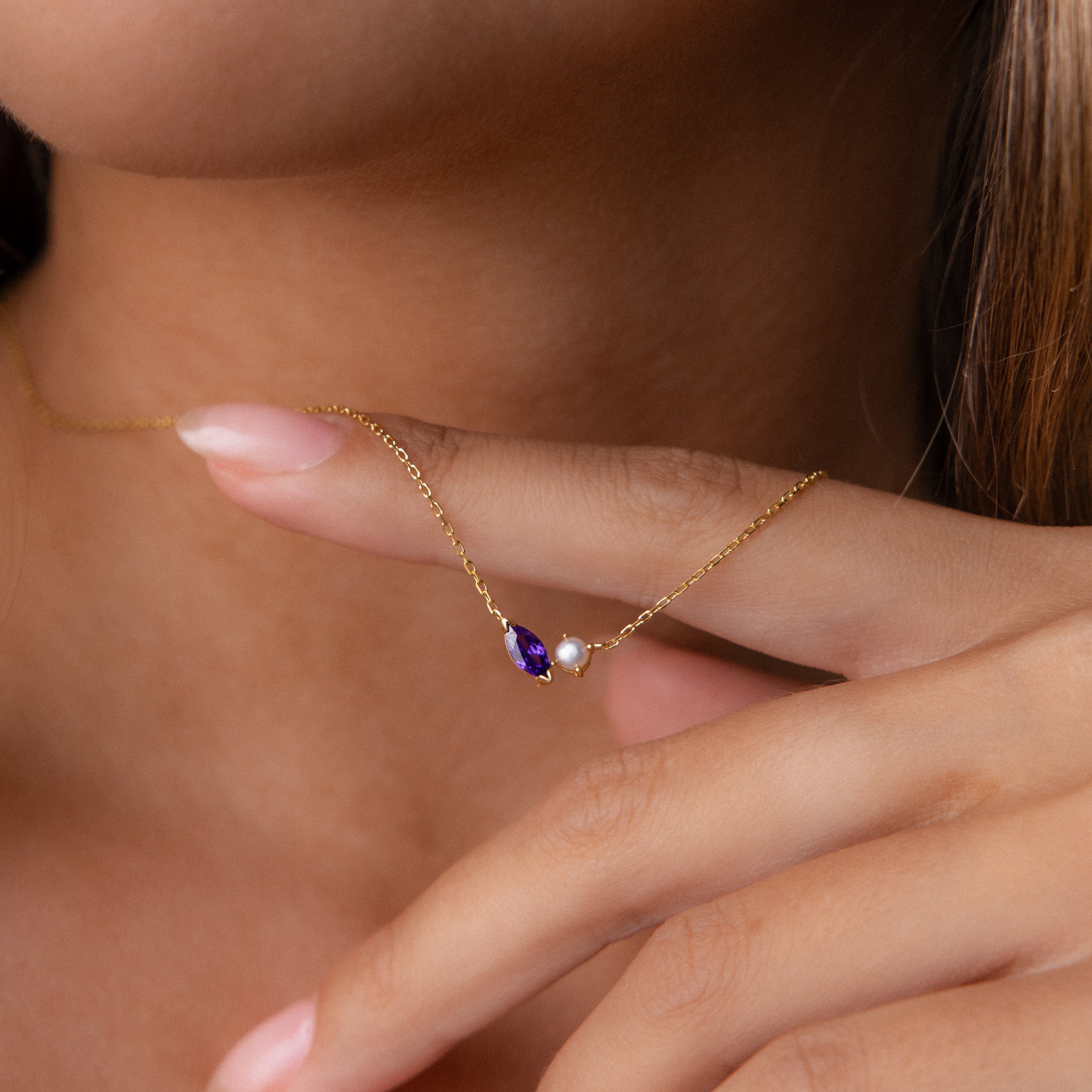 Close-up of a hand holding the Marquise Birthstone & Pearl Necklace, a gold customizable piece featuring a purple gem and a small pearl, shown against skin.
