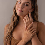 A woman with long hair smiles softly, touching her face as she wears a gold Marquise Birthstone & Pearl Necklace.