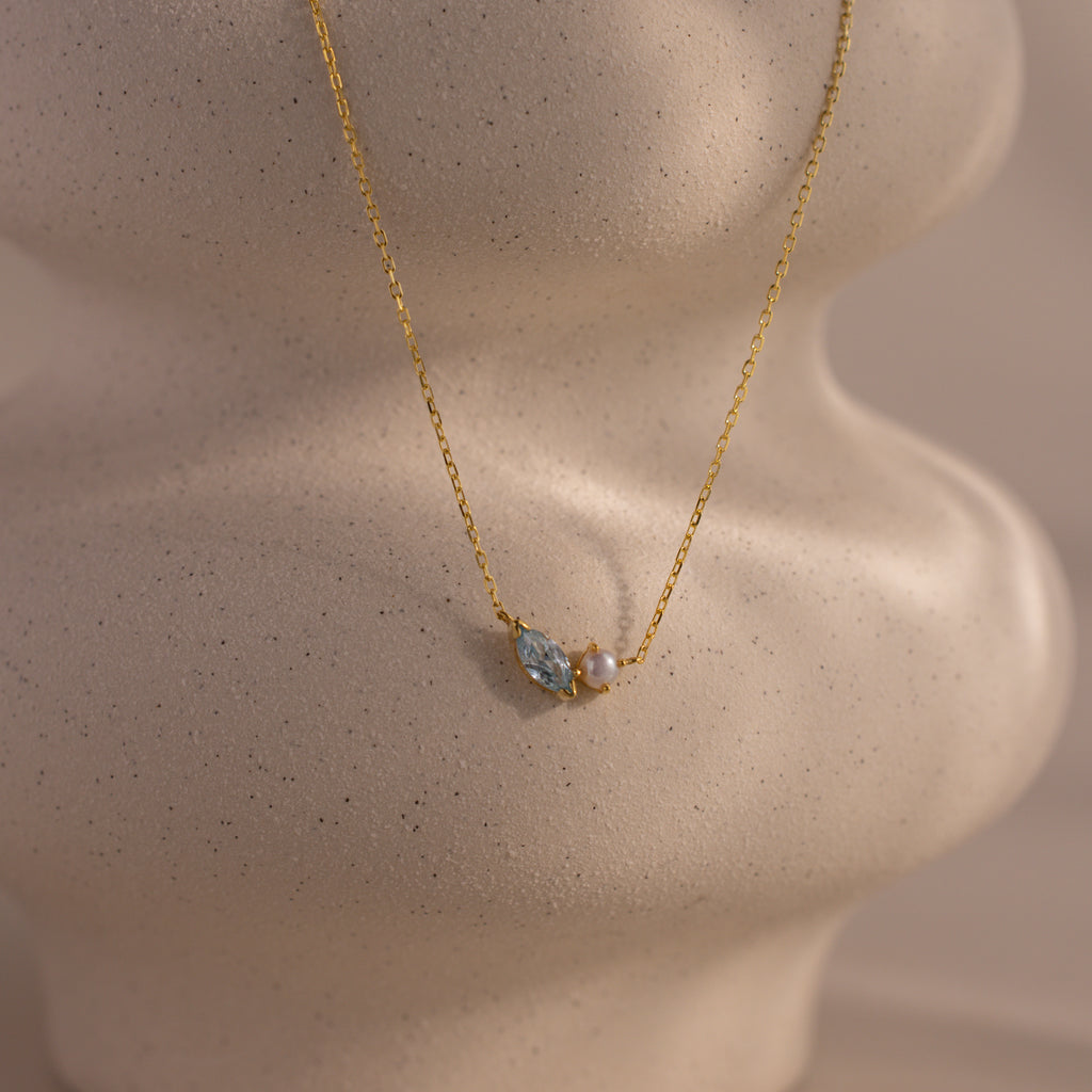 The Marquise Aquamarine & Pearl Necklace features a blue gemstone and pearl pendant, elegantly showcased on a textured beige mannequin bust.