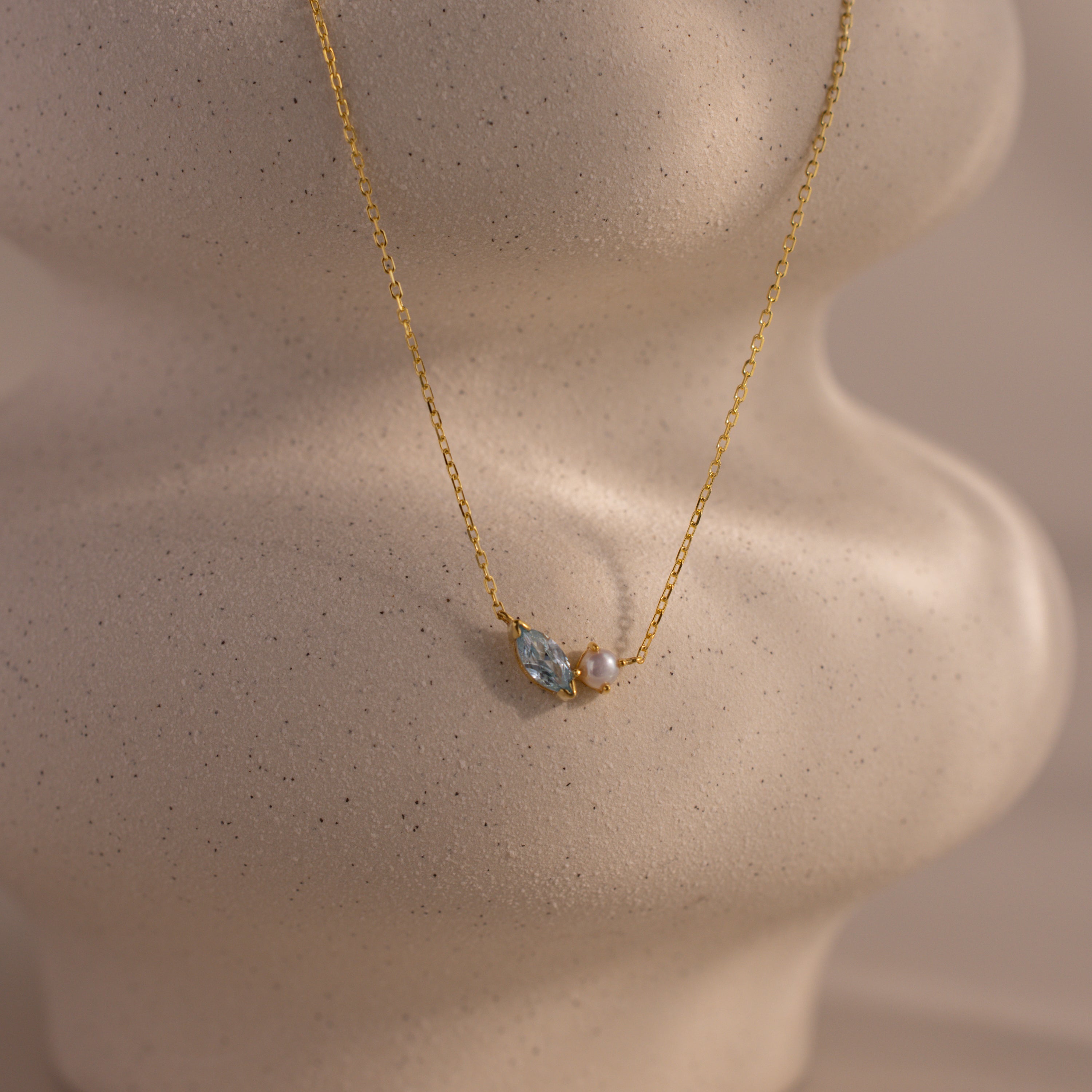 The Marquise Aquamarine & Pearl Necklace features a blue gemstone and pearl pendant, elegantly showcased on a textured beige mannequin bust.