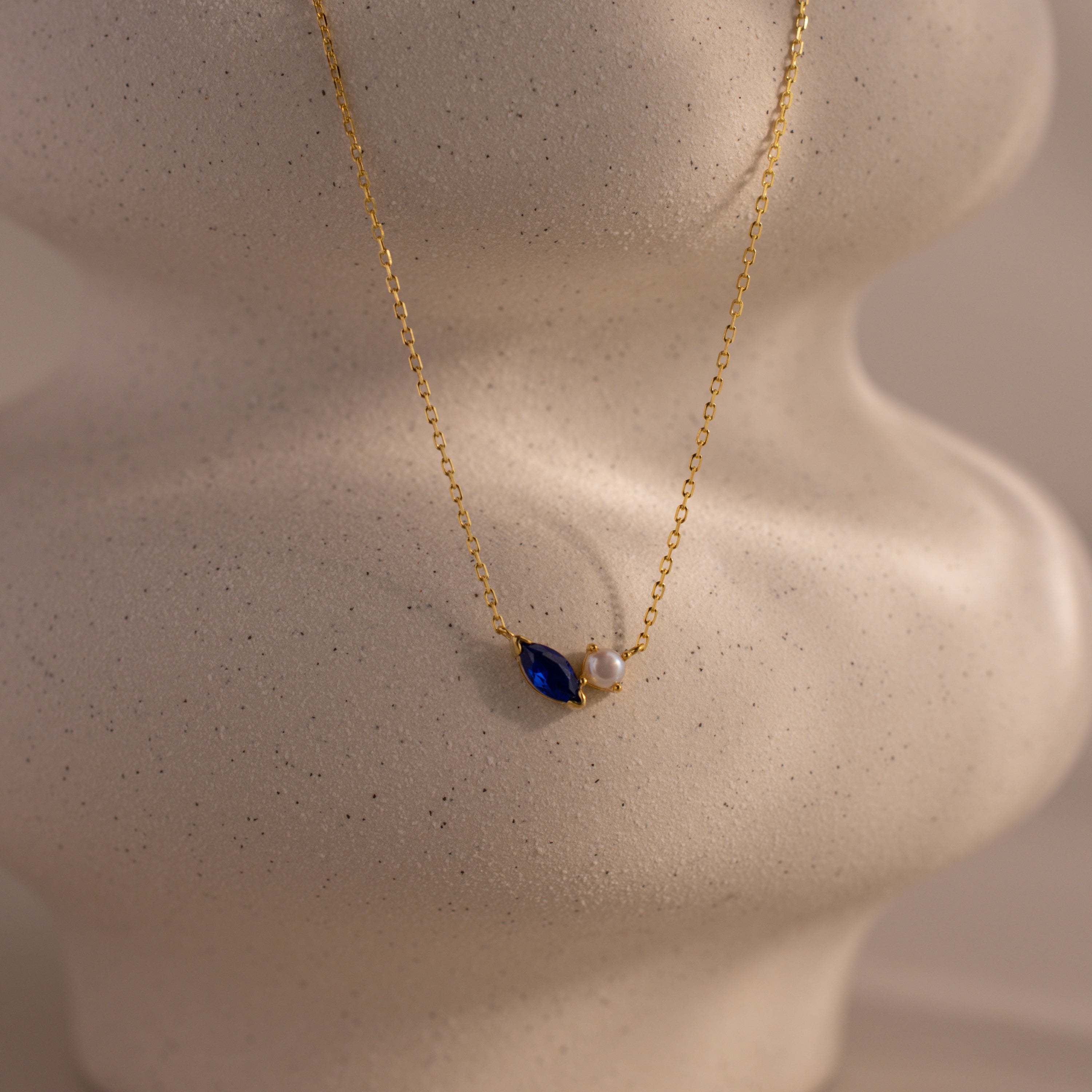 The Marquise Sapphire & Pearl Necklace features a blue marquise sapphire and a pearl, elegantly showcased on a neutral, textured bust.
