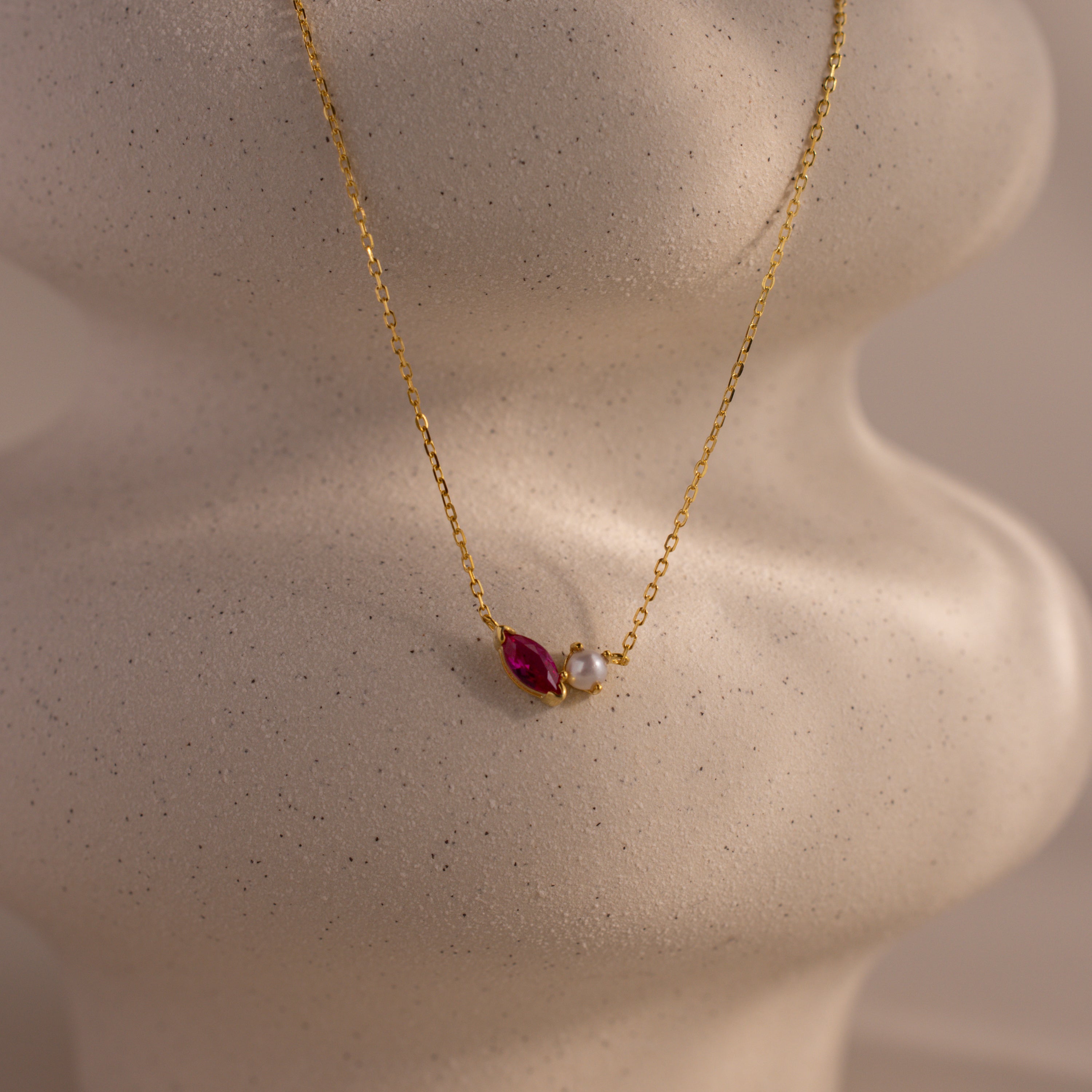 The Marquise Ruby & Pearl Necklace features a small red gemstone and pearl pendant, elegantly displayed on a cream-colored textured bust.