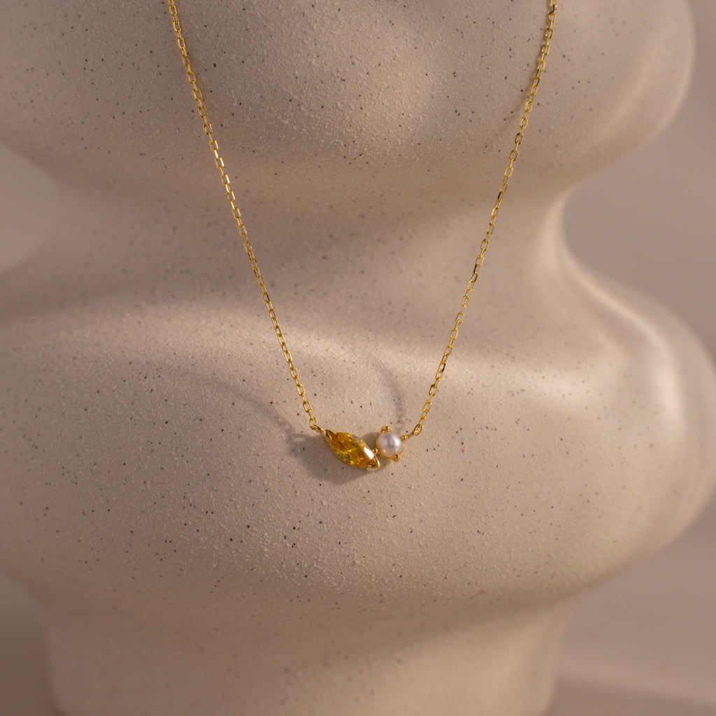 The Marquise Topaz & Pearl Necklace features a gold chain with a small leaf charm and pearl pendant, displayed on a textured light surface. This elegant necklace can be customized as a unique birthstone piece for a personal touch.