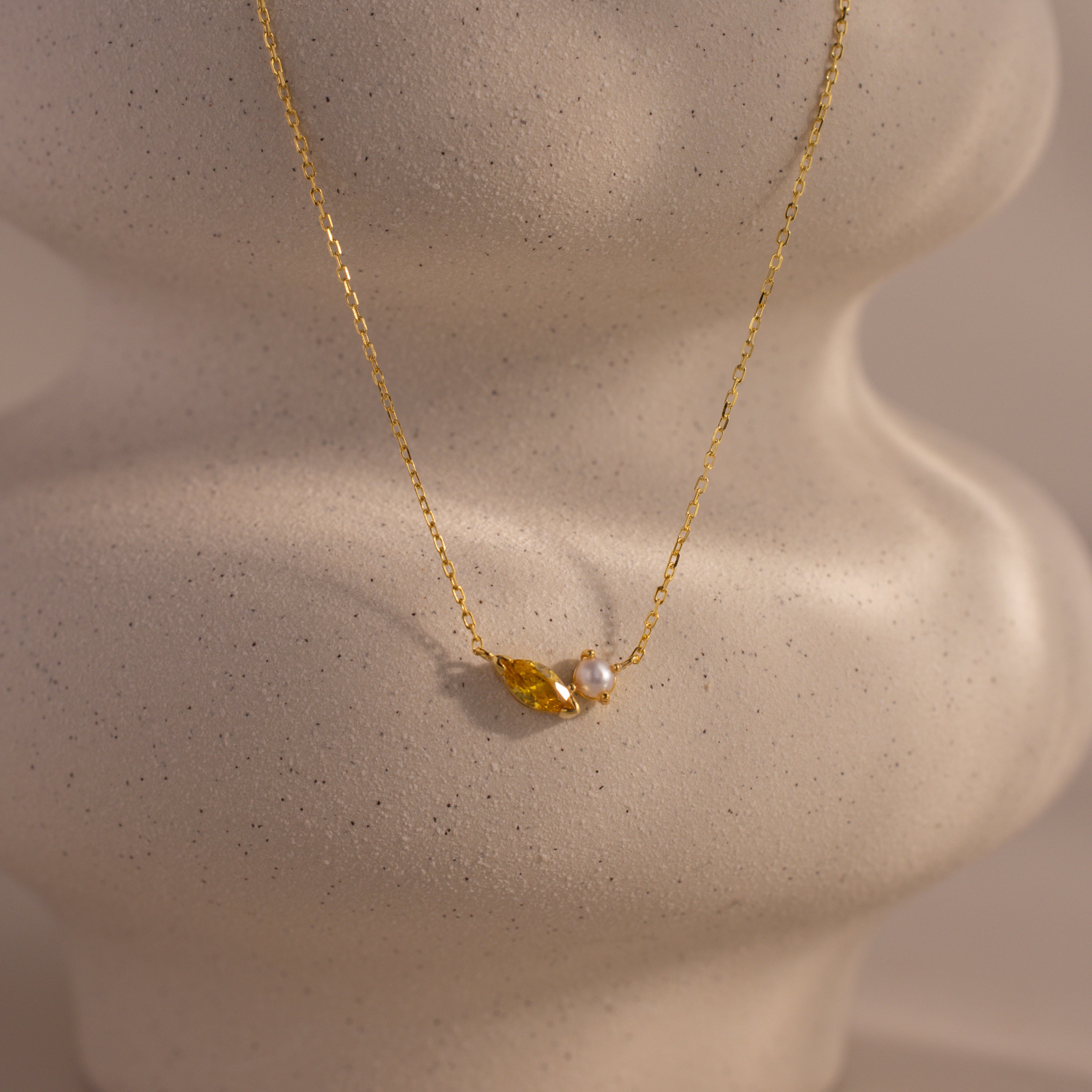 The Marquise Topaz & Pearl Necklace features a gold chain with a small leaf charm and pearl pendant, displayed on a textured light surface. This elegant necklace can be customized as a unique birthstone piece for a personal touch.