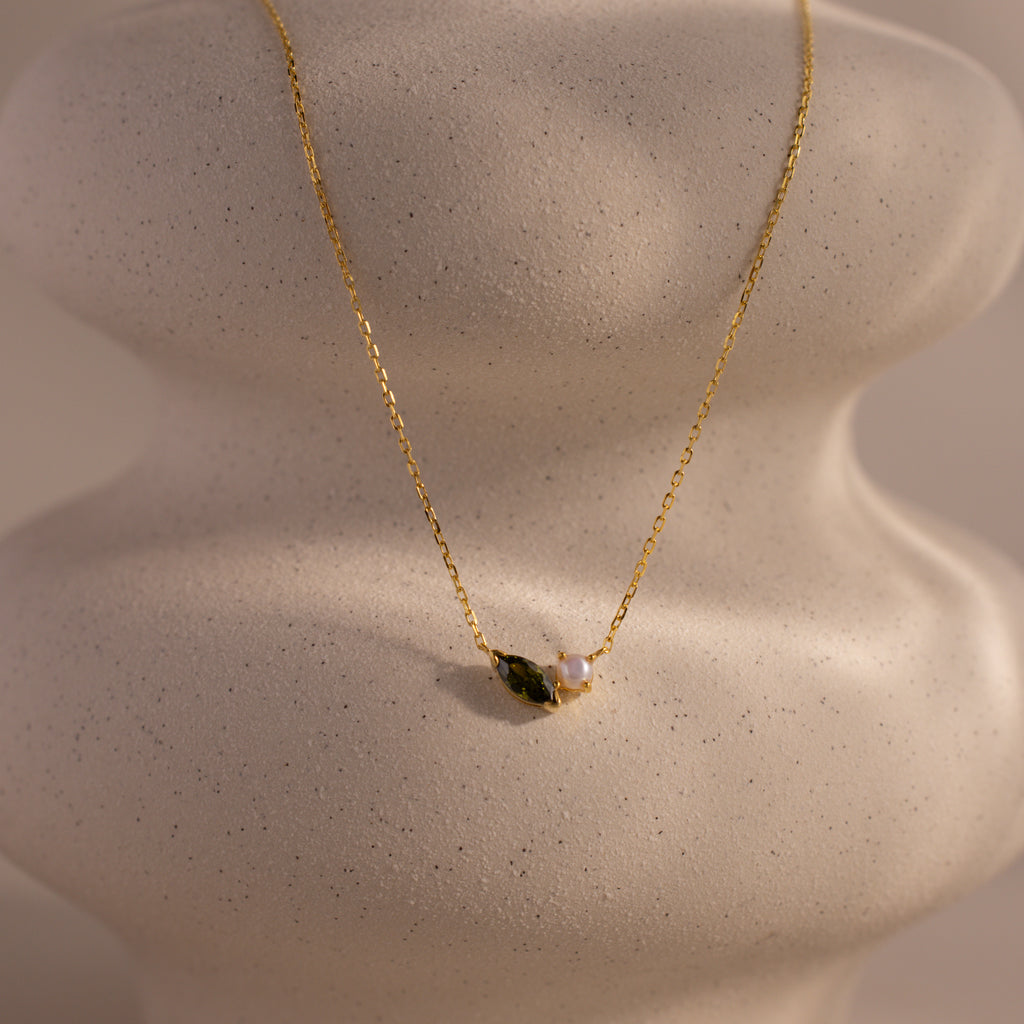 The Marquise Peridot & Pearl Necklace, featuring a small green marquise stone and pearl pendant, is elegantly displayed on a textured white jewelry bust.