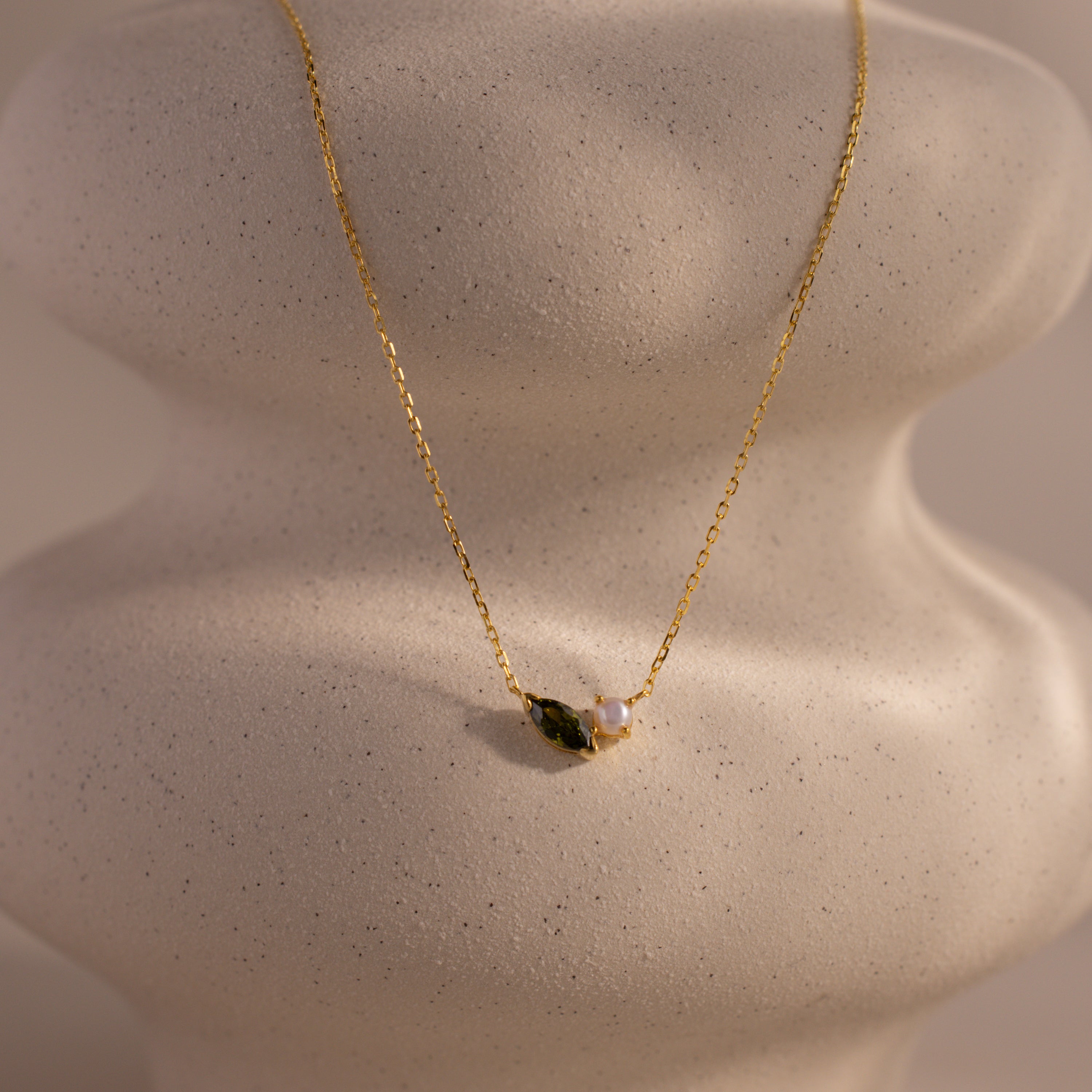 The Marquise Peridot & Pearl Necklace, featuring a small green marquise stone and pearl pendant, is elegantly displayed on a textured white jewelry bust.