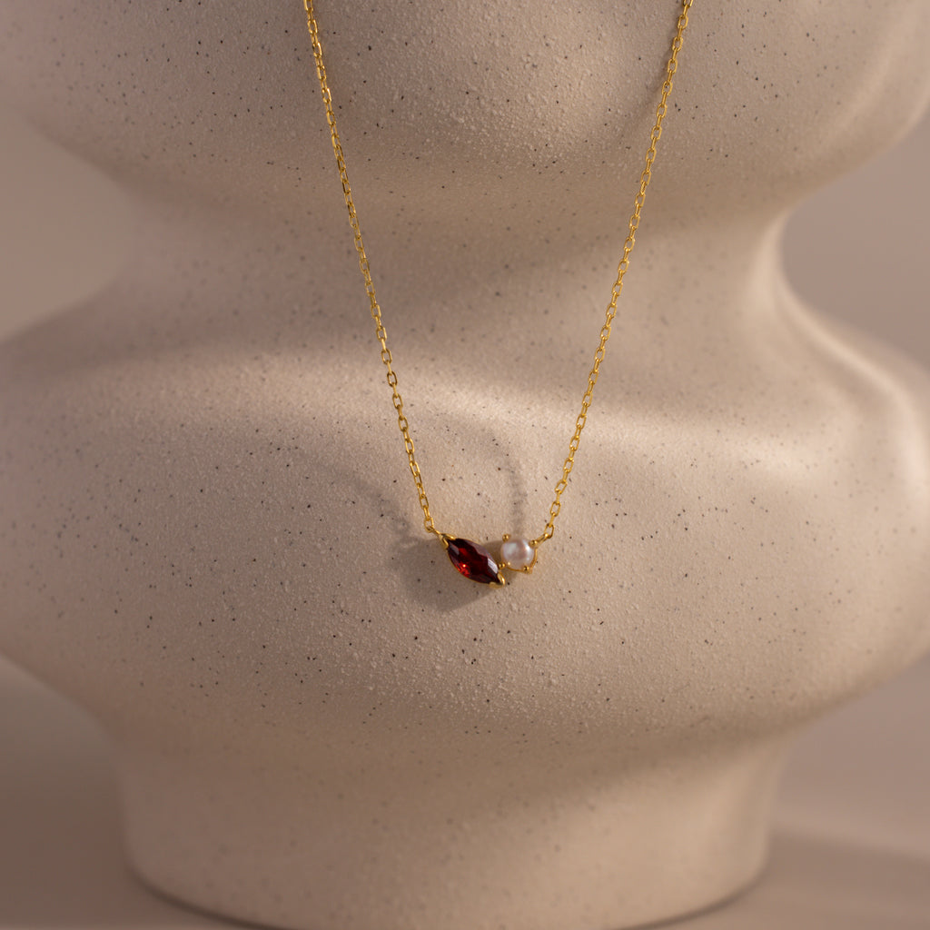 The Marquise Garnet & Pearl Necklace features a marquise-shaped garnet birthstone and a small pearl pendant, elegantly displayed on a textured beige bust.