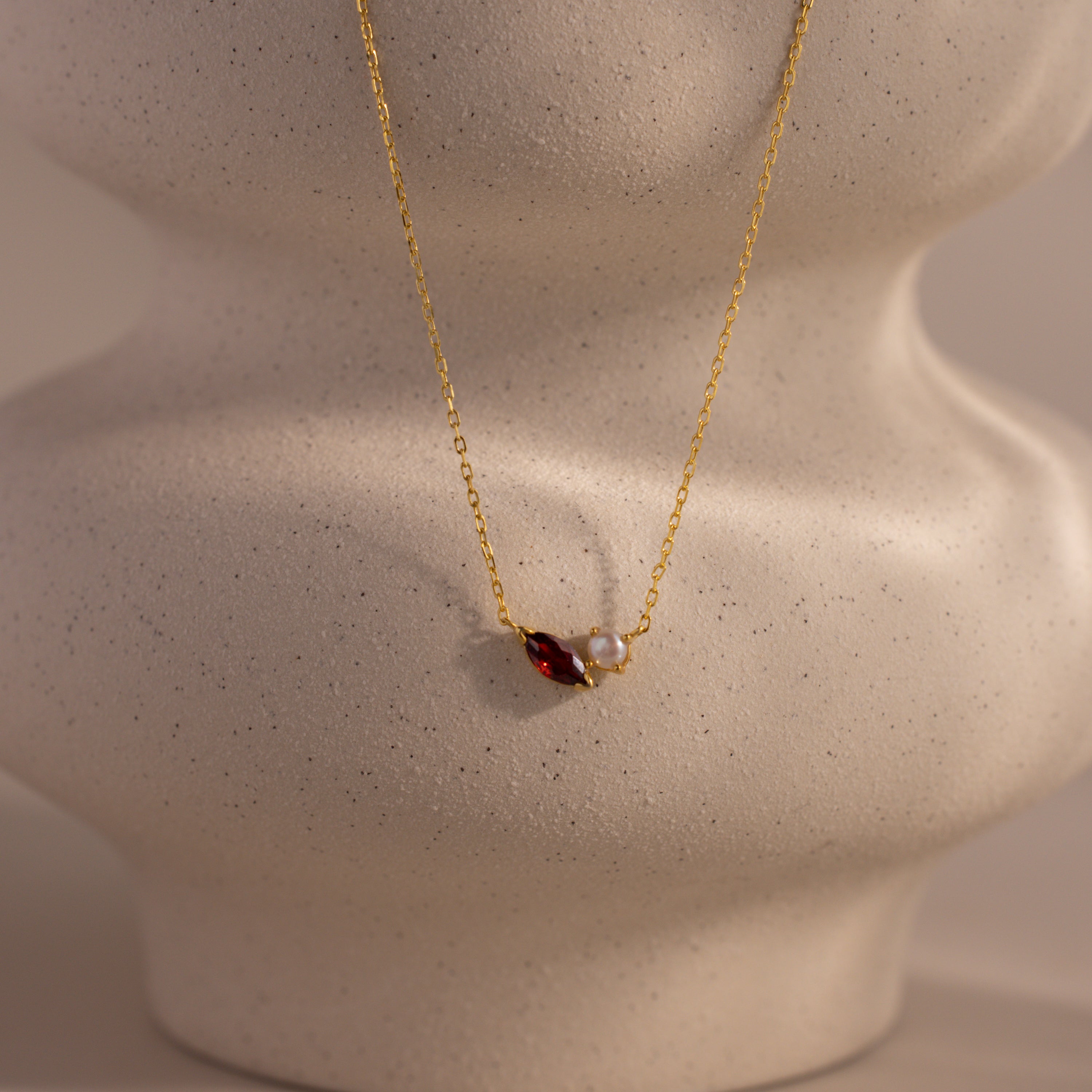 The Marquise Garnet & Pearl Necklace features a marquise-shaped garnet birthstone and a small pearl pendant, elegantly displayed on a textured beige bust.