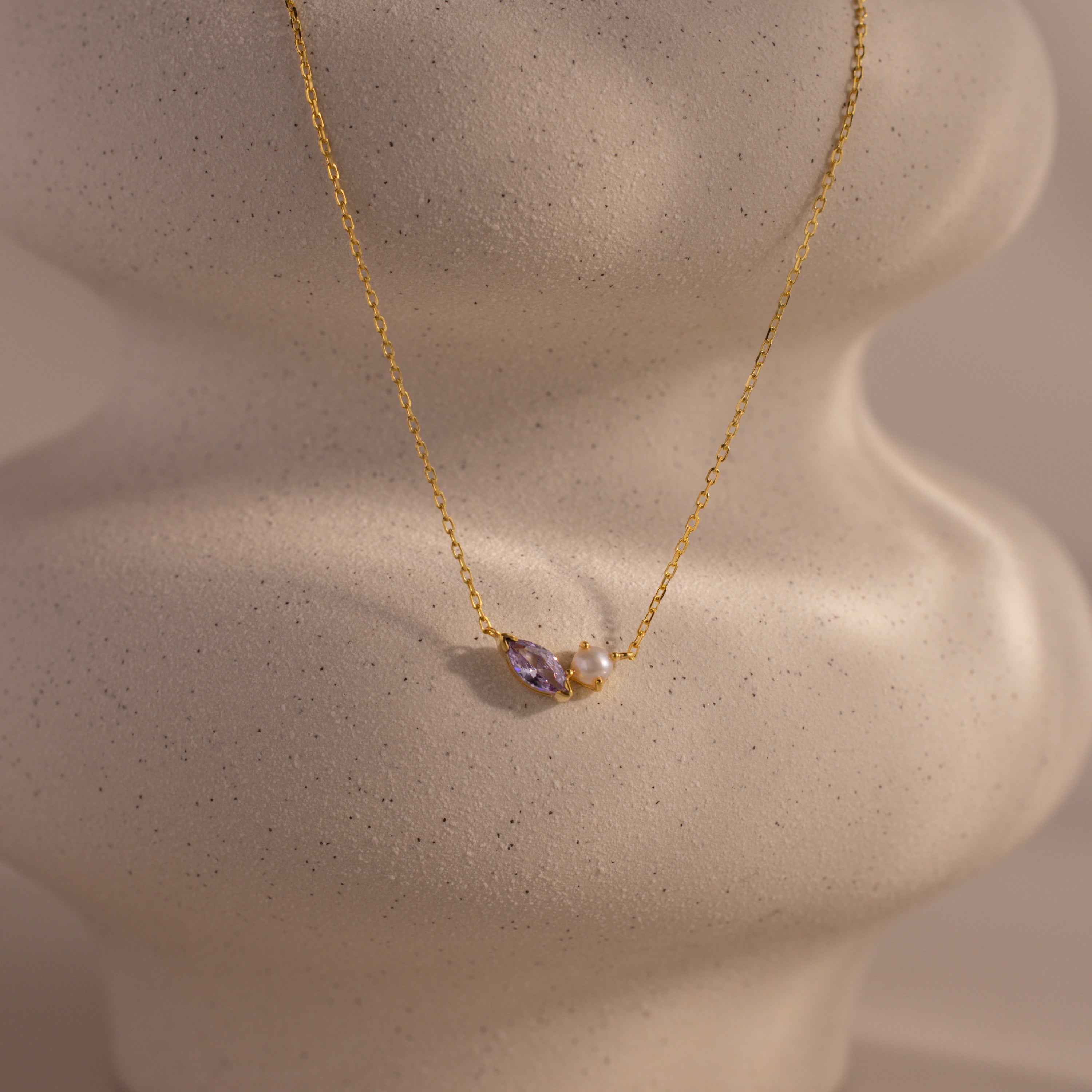 The Marquise Alexandrite & Pearl Necklace features a marquise-cut purple alexandrite and a pearl, displayed on a textured, wavy cream-colored stand.