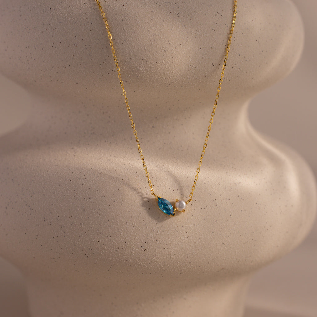 The Marquise Zircon & Pearl Necklace showcases a blue zircon teardrop and white pearl pendant on wavy light ceramic, making it an elegant, customizable birthstone necklace for a personal touch.