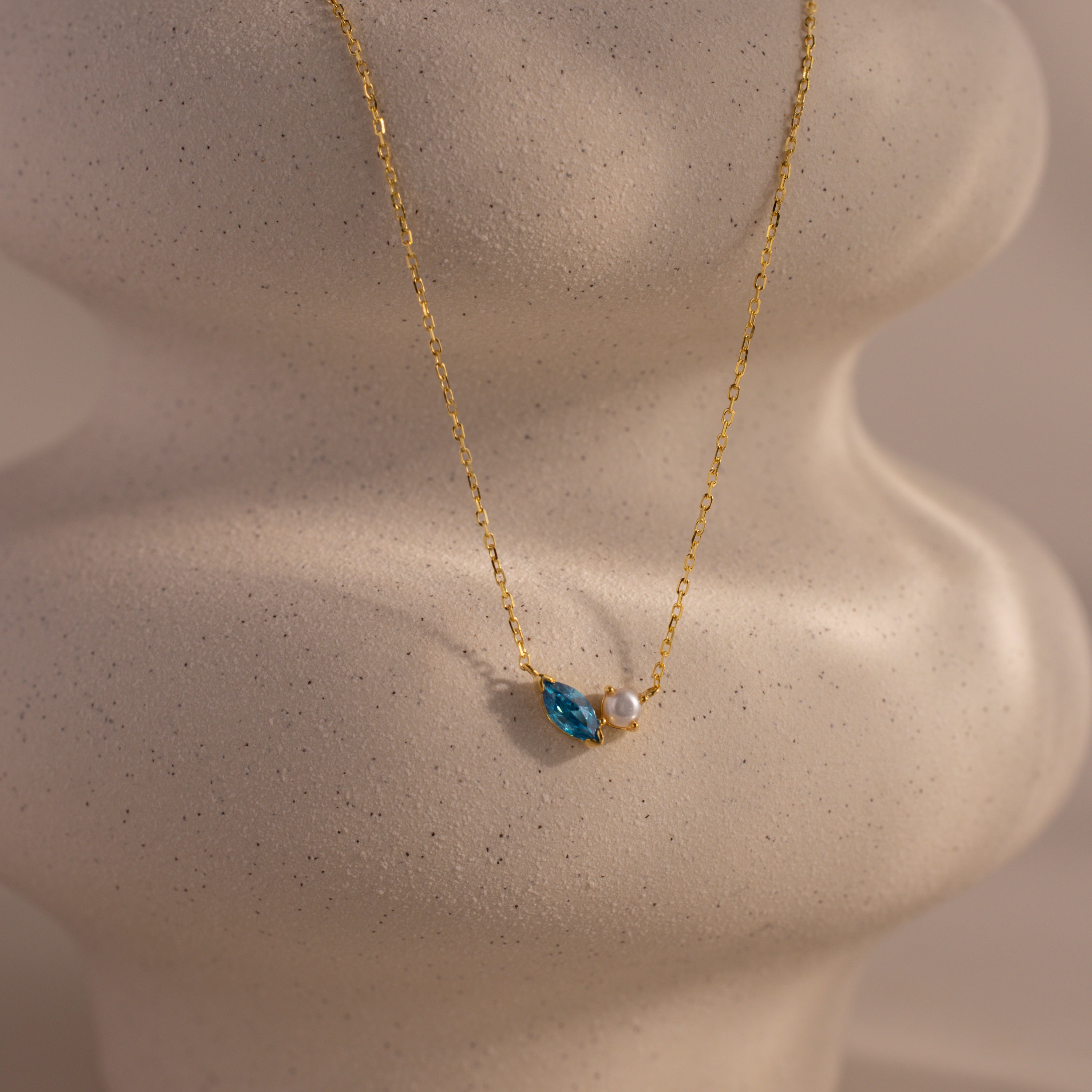 The Marquise Zircon & Pearl Necklace showcases a blue zircon teardrop and white pearl pendant on wavy light ceramic, making it an elegant, customizable birthstone necklace for a personal touch.