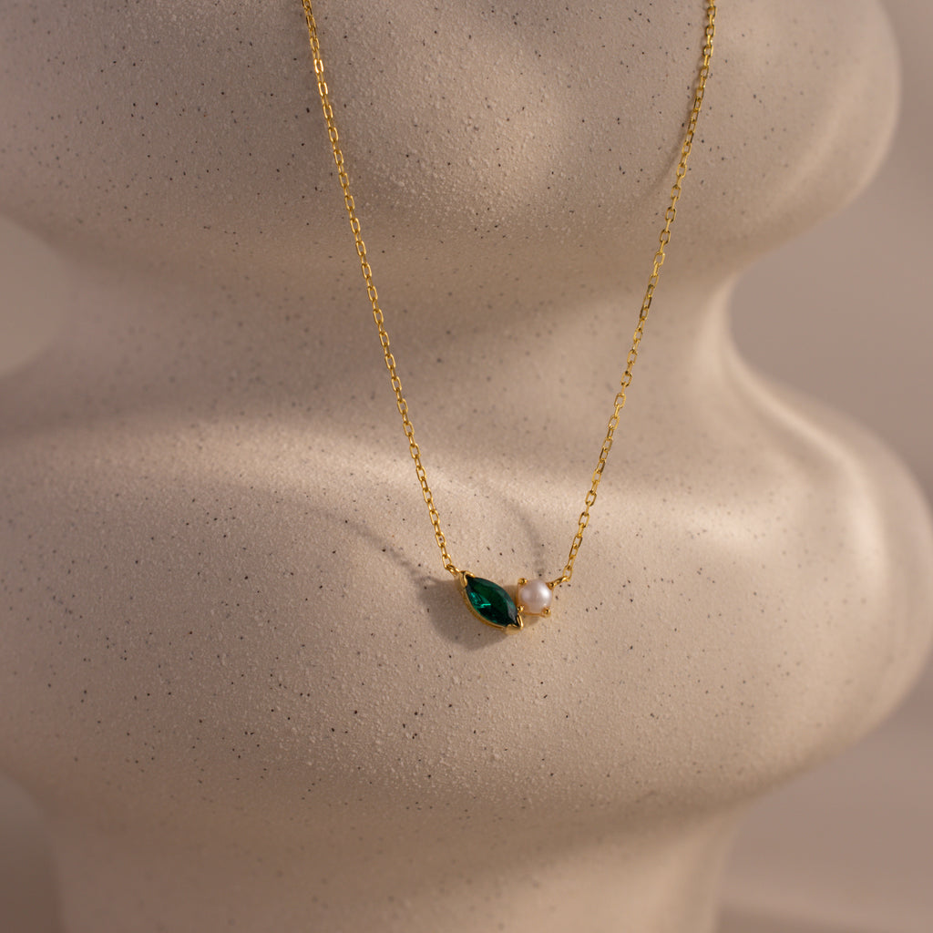 The Marquise Emerald & Pearl Necklace features a green marquise emerald birthstone and a delicate pearl pendant, displayed on a textured, sculpted surface.