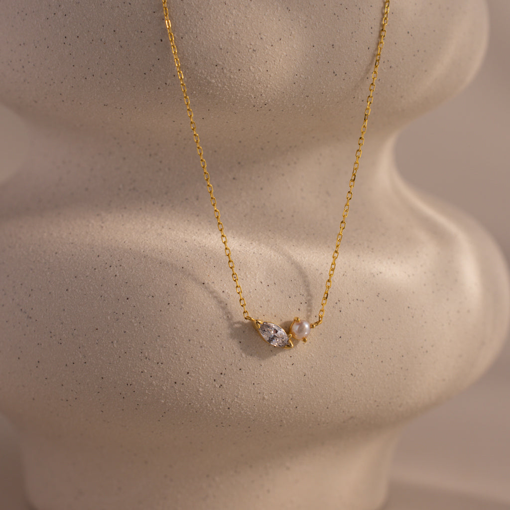 The Marquise Diamond & Pearl Necklace features a gold chain with pearls and a marquise diamond pendant, elegantly displayed on a textured beige background. Wear it solo or layered—perfect for adding customizable style to any outfit.