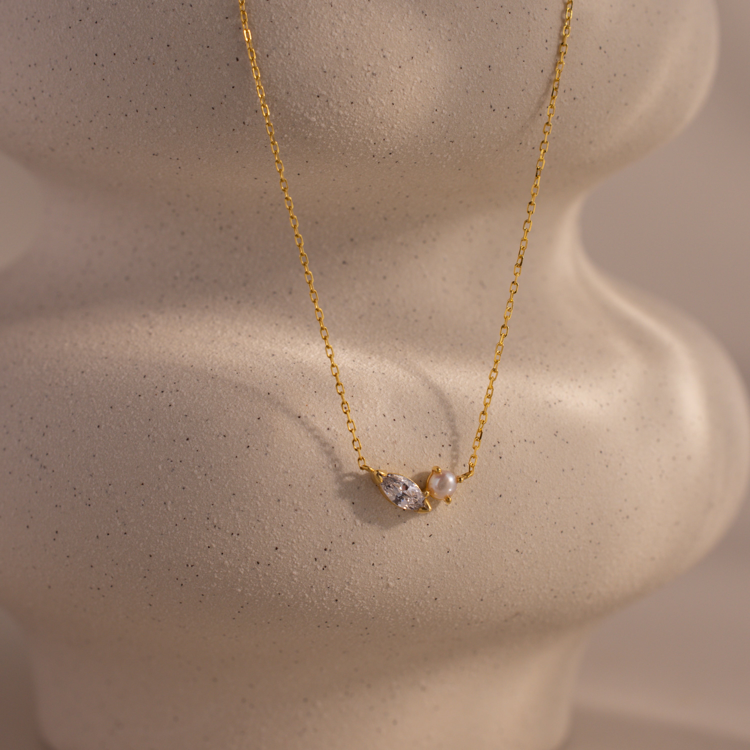 The Marquise Diamond & Pearl Necklace features a gold chain with pearls and a marquise diamond pendant, elegantly displayed on a textured beige background. Wear it solo or layered—perfect for adding customizable style to any outfit.