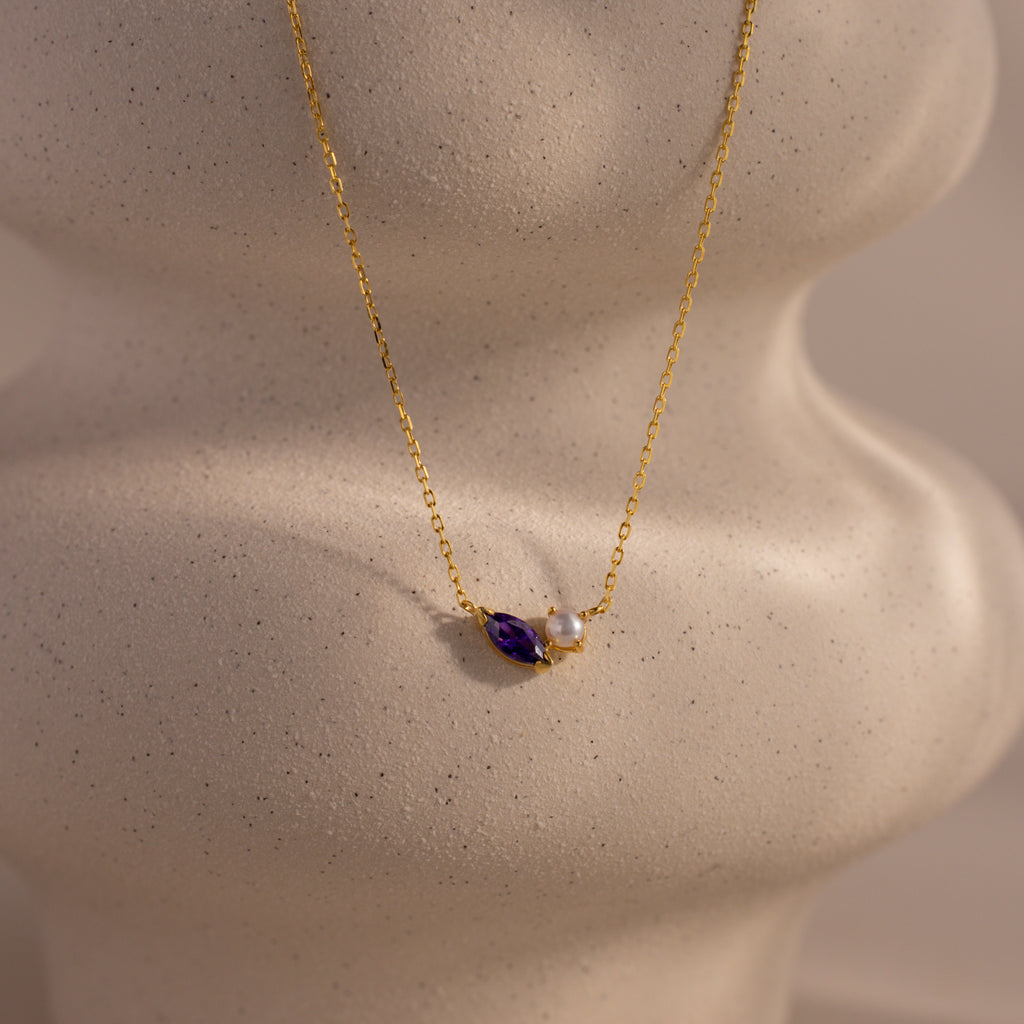 The Marquise Amethyst & Pearl Necklace is a gold piece featuring a purple marquise amethyst birthstone and a pearl pendant, shown on a white, textured display bust.