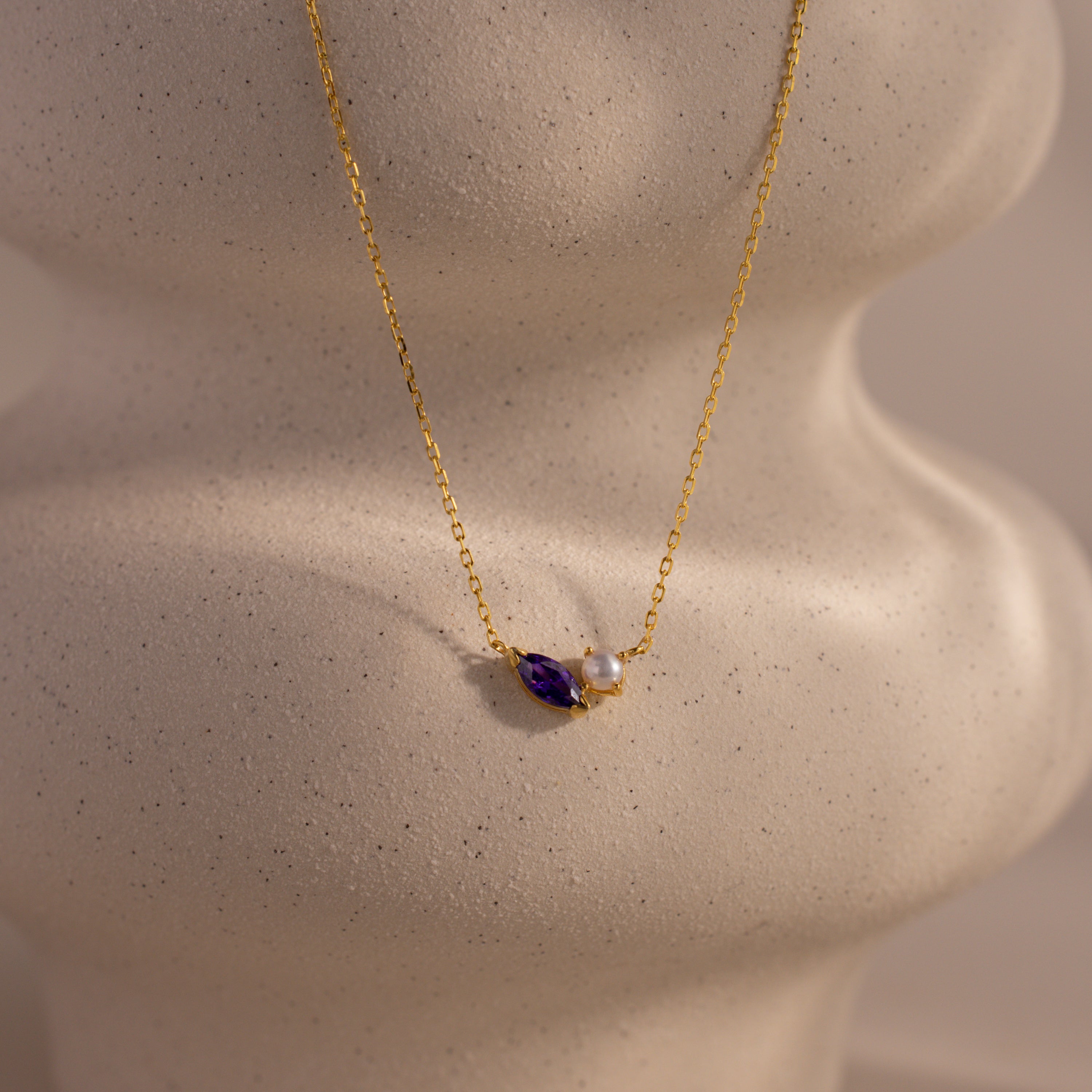 The Marquise Amethyst & Pearl Necklace is a gold piece featuring a purple marquise amethyst birthstone and a pearl pendant, shown on a white, textured display bust.