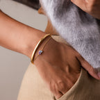 A hand in a pocket wears a gold bangle and the Toi et Moi Teardrop Heart Bracelet in 18K Gold, featuring a purple gemstone charm for timeless elegance.