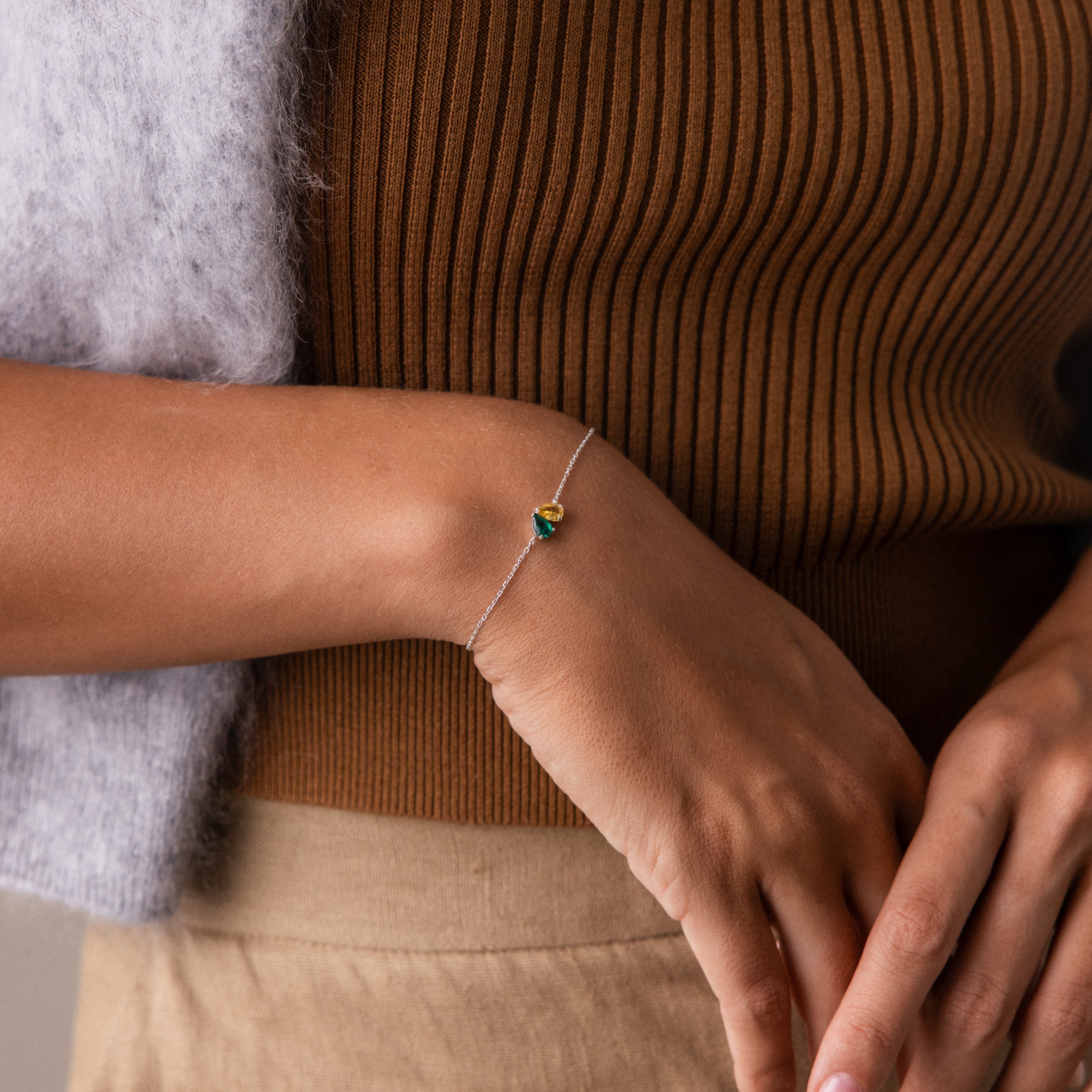 Close-up of a woman's hand wearing the Toi et Moi Teardrop Heart Bracelet in Sterling Silver, featuring a green gemstone, with a brown ribbed top in the background.