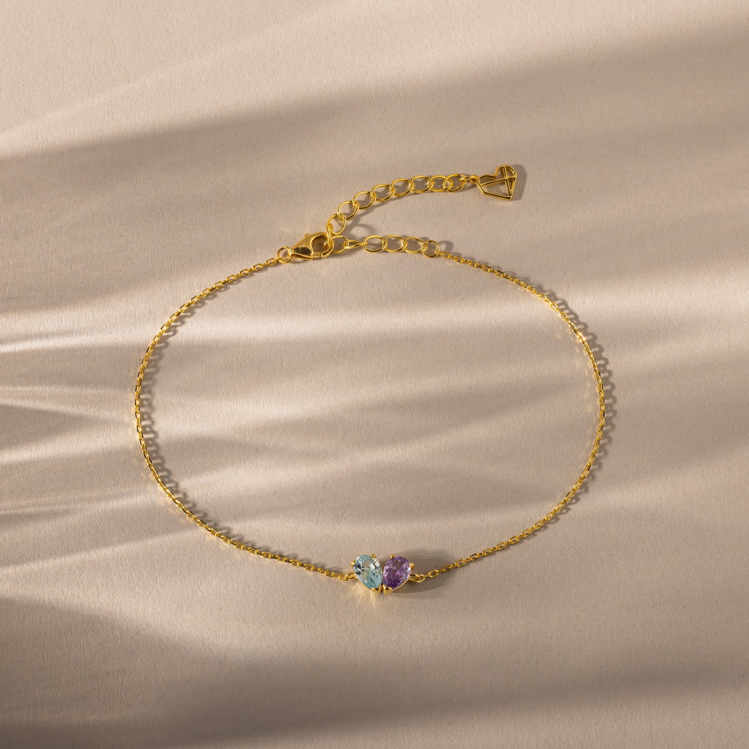 The Toi et Moi Teardrop Heart Bracelet in 18K Gold features three colorful gemstones, including pear-cut stones, elegantly displayed on a beige surface with soft shadows.