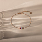 The Toi et Moi Teardrop Heart Bracelet in Rose Gold features a red and a clear gemstone, elegantly displayed on a beige surface with soft shadows.