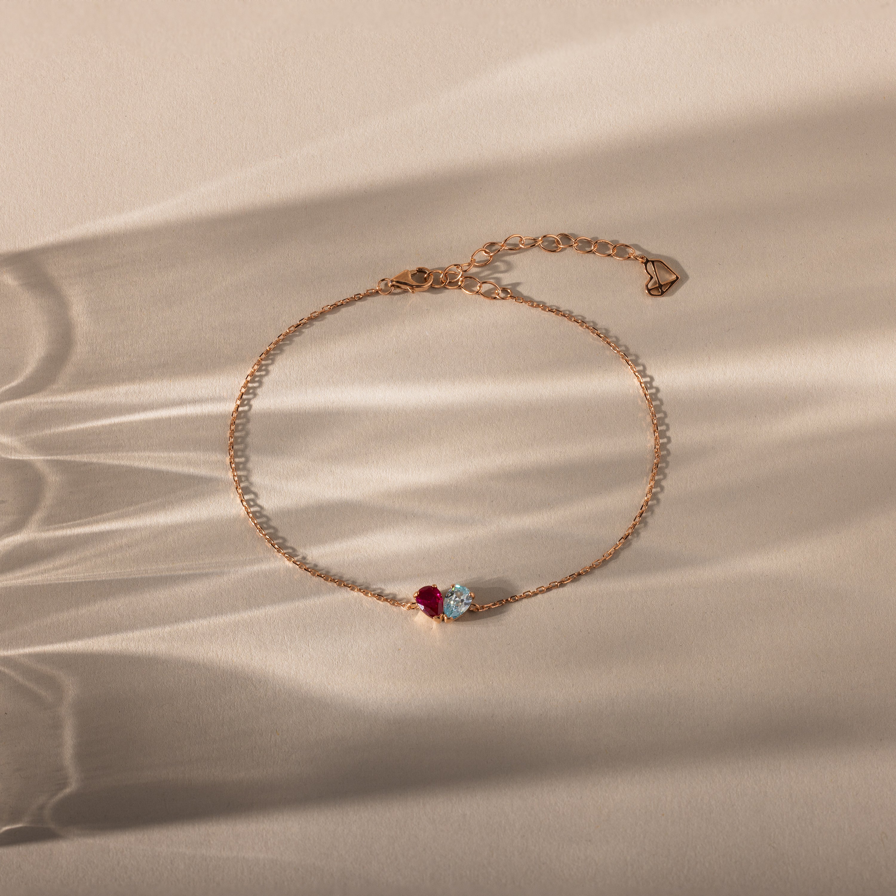 The Toi et Moi Teardrop Heart Bracelet in Rose Gold features a red and a clear gemstone, elegantly displayed on a beige surface with soft shadows.