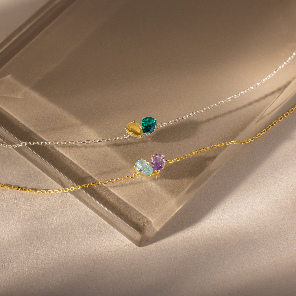 The Toi et Moi Teardrop Heart Bracelet in Sterling Silver, featuring colorful gemstone heart charms, rests alongside another delicate chain bracelet on a sunlit, neutral surface.
